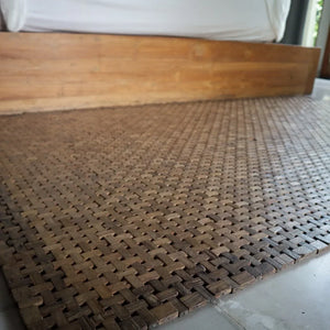 Handwoven wooden mat on floor beside wooden bed, eco-friendly home decor accent