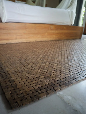 Handwoven wooden mat on floor beside wooden bed, eco-friendly home decor accent
